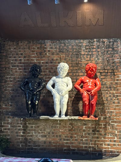 The image displays an outdoor scene in Dongguan, China, featuring three "Manneken Pis"-style statues mounted against a reddish-brown brick wall. Above the wall, a dark brown facade features the raised metallic letters "ALIKIM", illuminated by an overhead light source.
The three statues are arranged horizontally on a narrow ledge. From left to right, they are: one glossy black, one matte white, and one glossy red. Each statue depicts a nude, cherubic male child in a posture consistent with urination. Visible streams of liquid are actively emanating from the genital area of each statue, flowing downwards onto the brickwork below. These streams are clear against the darker brick and cast faint shadows.
In the lower foreground, a red and white checkered surface, likely a tablecloth, is partially visible. A dark, rounded object, possibly a pot or container, rests on this surface. The overall lighting suggests an evening or nighttime setting, with artificial illumination highlighting the statues and the "ALIKIM" sign. No human individuals are present in the frame.