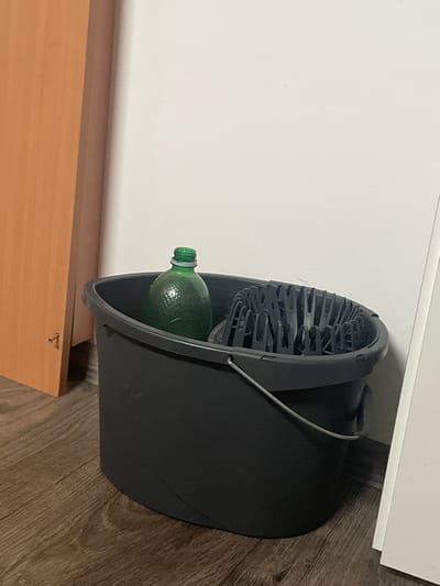In a room in Teplice, Czechia, a dark gray bucket with a silver handle sits on a wooden floor. Inside the bucket, a green glass bottle with a ribbed texture is visible, along with a black plastic mop wringer. To the left of the bucket is a section of a light brown wooden door or cabinet, and to the right, a portion of a white wall is present.