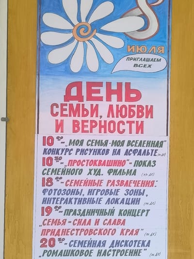 The image shows a poster on a yellow wall, likely in Tiraspol City Council, Moldova. The poster features a large daisy graphic at the top and colorful text announcing events. The main heading in large red letters reads "ДЕНЬ СЕМЬИ, ЛЮБВИ И ВЕРНОСТИ" (Day of Family, Love and Fidelity). Below this, a list of events with dates and times is presented. These include a drawing contest on asphalt on July 10th at 10:00, a film screening on July 10th at 10:30, and family entertainment with photo zones, game areas, and interactive locations on July 18th. On July 19th, there's a concert titled "Family is Strength and Glory," and on July 20th at 20:30, a family disco with the theme "Daisy Mood" is scheduled. The text is handwritten in Russian.