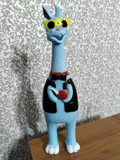 The image shows a blue, anthropomorphic toy figure with a long neck, large yellow sunglasses, a red bowtie, a black vest, and it is holding a red pipe. The figure is standing on a wooden surface, with a patterned wallpaper in the background. The figure appears to be a novelty item or a collectible. There are no people or any other elements visible that would indicate a specific location in Luhansk, Ukraine, or any other city. The scene is a simple still life composition.