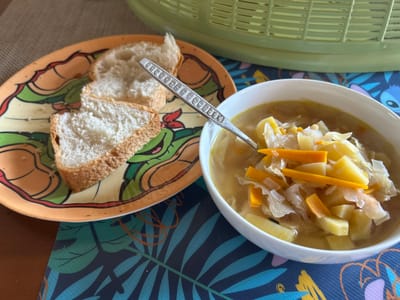 A white bowl of vegetable soup, containing potatoes, carrots, and cabbage, sits on a dark blue placemat with a tropical leaf pattern. To the left of the soup bowl is a colorful plate with cartoon characters, holding three slices of white bread. A spoon with a patterned handle rests in the soup. In the background, a green colander is visible. The image appears to be taken indoors, likely in a home environment in Dehiwala, Sri Lanka. There are no people visible in the image.