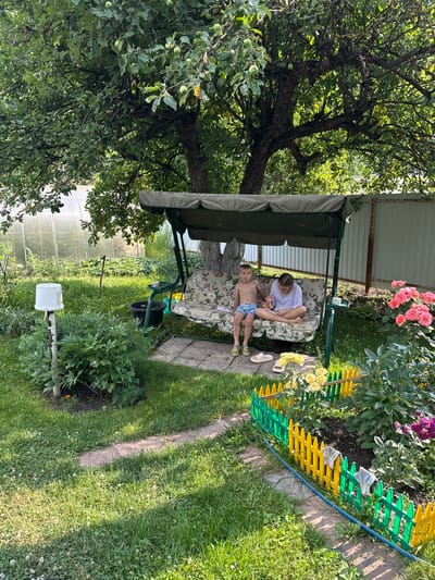 A young boy and a girl are relaxing on a garden swing. The boy, wearing blue swim trunks, is lying down on the swing, facing the camera. The girl, wearing a white t-shirt, is sitting up and appears to be reading a book or looking at her phone. They are in a backyard setting with lush green grass and a variety of plants and flowers, including pink and yellow roses, and dark pink peonies. A large tree with green leaves and some small, unripe apples is to the left of the swing. In the background, a greenhouse structure is visible. The scene suggests a warm, sunny day, likely in the summer, given the attire of the children and the blooming flowers. The overall mood appears relaxed and peaceful, indicative of a leisurely afternoon outdoors.