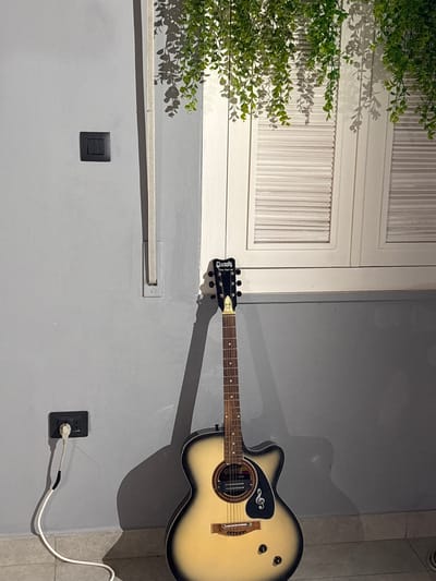 In the city of Sweileh, Jordan, an acoustic guitar stands upright against a light gray wall. The guitar has a light-colored body with black trim and a dark fretboard. A black electrical outlet with a white cord plugged into it is mounted on the lower portion of the wall to the left of the guitar. A second, black electrical switch is located higher up on the wall, also to the left of the guitar. Above the guitar and to the right, a white shuttered window is visible. Hanging greenery drapes down from the top of the frame, casting shadows on the wall and window. The floor appears to be tiled. The scene is lit with artificial light, casting a prominent shadow of the guitar onto the wall.
