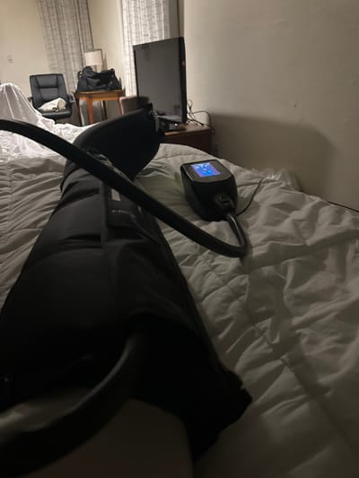 The media file captures an indoor scene, likely a bedroom, illuminated by dim lighting that suggests it is evening or night. A person's legs are visible, with one leg prominently in the foreground, extending across a bed with white, rumpled sheets. This leg is encased in a black compression boot, part of a therapeutic or recovery system.
On the bed, near the person's upper thigh, rests a compact, black control unit or pump connected to the leg boot by a dark hose. This device features a small, illuminated blue screen displaying digital information. Visible text on the screen includes "OFF" along with other indistinct symbols or numbers, indicating the device's operational status.
In the background, to the left, a flat-screen television sits on a dark stand. A piece of wooden furniture, possibly a bedside table or dresser, is partially visible next to the TV. Further to the left, white window blinds or curtains are visible, suggesting a window. The overall ambiance is one of quiet rest or recuperation, with the person seemingly undergoing a leg compression treatment. The scene does not contain any explicit visual cues for the Richmond, United States location beyond the provided context.