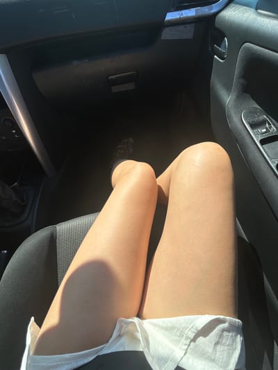 An overhead, high-angle shot shows a person's legs from the knees down, seated inside a vehicle. The person is wearing a white garment that covers their lap and is partially visible. The interior of the car is dark, with sunlight streaming in, casting shadows on the person's legs. The dashboard and a portion of the car door are visible, suggesting the photo was taken from the passenger seat, looking towards the driver's side. The person's feet are obscured by shadow, but one foot appears to be wearing a patterned sock or shoe. The car's interior appears to be a standard model, with a gear stick visible on the left side of the frame. The lighting suggests it is daytime. The location of Zagrađe, Montenegro, is not discernible from the visual information present in the image.