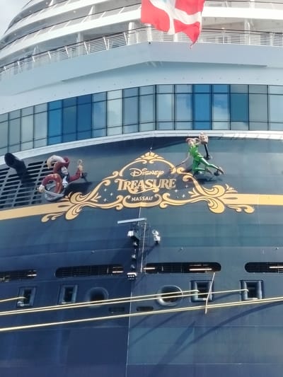 The image depicts the Disney Treasure cruise ship docked in Nassau, Bahamas, not San Miguel de Cozumel, Mexico. The ship features the "Disney Treasure NASSAU" logo with Captain Hook and Peter Pan figures adorning the hull. A Danish flag flutters atop the ship, and windows reflecting the sky are visible on the upper decks. Mooring lines are attached to the ship's side. The ship is dark blue, while the characters and logo are in white and gold.
