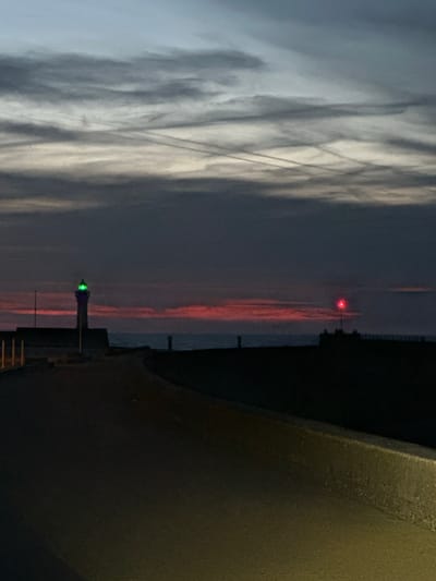 The image shows a twilight view of the harbor entrance in Saint-Valery-en-Caux, France. The sky is a gradient of dark blue at the top, transitioning to a band of reddish-orange clouds on the horizon, and then to a lighter gray-blue. Thin, linear contrails crisscross the upper sky.
In the middle ground, two navigation lights are visible. On the left, a lighthouse with a green light is positioned near a stone pier. On the right, a smaller structure with a red light is silhouetted against the sky. The sea is a dark, calm expanse between the land and the horizon.
In the foreground, a paved pathway curves from the bottom left towards the right of the image. The pathway appears to be made of asphalt or a similar dark material. A concrete barrier runs along the right edge of the pathway, with a textured surface. The lighting suggests it is either dawn or dusk, with artificial lights casting a faint glow on the pathway. No people are visible in the scene.