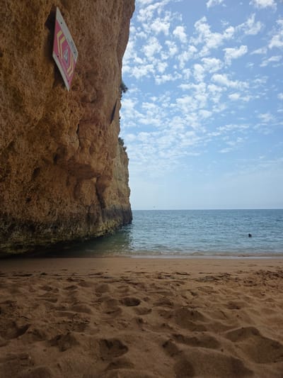 The image depicts a beach scene in Porches, Portugal. The composition is vertical, with the foreground on the left featuring a rocky cliff face and a sign. The cliff is a warm, sandy brown, textured with rough surfaces and crevices, indicative of natural erosion. A triangular sign with a pink and yellow design is affixed to the rock face. It appears to have text and an image, possibly a warning or informational notice.

The middle ground shows the transition from the rocky shore to the ocean and beach. The water is a tranquil blue-green, with gentle waves lapping at the shore. A dark object, possibly a piece of debris or a small animal, is visible in the water.

The background is dominated by a clear blue sky with scattered, fluffy white clouds. The right side of the image is the sandy beach, characterized by its fine, light brown grains and visible footprints. The horizon line where the sky meets the sea is sharp and distant, suggesting an open, expansive view.

The lighting suggests it is daytime, likely late morning or early afternoon, given the brightness of the sky and the clear shadows. The weather appears pleasant, with calm seas and scattered clouds indicating fair conditions. The overall mood is serene and natural, evoking a sense of peace and the beauty of the coastal landscape. The location cues like the specific rock formations and the typical beach setting are consistent with a Portuguese coastal town like Porches.