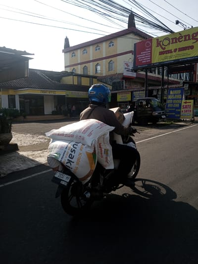A person on a motorcycle laden with large sacks is riding on a road in an urban environment. The rider wears a blue helmet and is transporting several white sacks, some marked with "USAKI Gula Kristal Putih." The motorcycle's license plate reads "F 2014 MP." In the background, there is a building with a sign saying "FRONT OFFICE Bubur Ayam." Across the street, there are several food vendors and signs for "Konèng HOTEL & RESTO," "BAKSO MALANG," and "SATE & SOTO PADANG." The surroundings indicate a busy commercial area with overhead power lines and a mix of buildings.