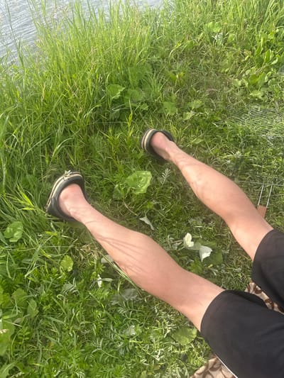 The image shows a person's legs and feet resting on a grassy area near a body of water. The person is wearing black shorts and black and yellow slip-on sandals. The grass is green and appears to be growing along the edge of the water. There is a metal grill in the background to the right. The sun is shining, casting shadows on the legs, indicating it is daytime. The overall scene suggests a relaxed outdoor activity, possibly a picnic or leisure time by the water. The location is Ozyorny, Russia.