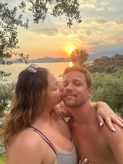 A candid, close-up, chest-up, outdoor, selfie-style photograph captures a couple in an embrace during sunset. The woman, on the left, is kissing the man on the cheek. She has long, wavy brown hair, and sunglasses perched on her head. She is wearing a gray tank top with maroon trim. The man, on the right, has short brown hair and a light beard. He is shirtless and wearing a small stud earring in his left ear. A woman's hand, with white nail polish, rests on his shoulder, and she is wearing a beaded bracelet on her wrist.
The background features a serene seascape with calm water reflecting the warm hues of the setting sun. Distant mountains form a silhouette against the sky, which is filled with dramatic clouds illuminated by the golden sunlight. Olive trees and lush green foliage are visible in the mid-ground, suggesting a Mediterranean setting. The location is the city of Garda, Italy, on the shores of Lake Garda, known for its picturesque landscapes and olive groves.