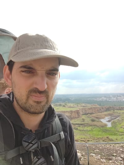 An adult male is prominently featured in the foreground of a selfie-style photograph taken from an elevated outdoor position in Rosh Haayin, Israel. He wears a light-colored baseball cap and a dark hooded sweatshirt, with sunglasses hanging from his chest strap. A dark backpack is visible on his shoulders. A portion of a second individual's head, possibly a child, is partially visible behind and to the left of the main subject's head. The background reveals a natural landscape encompassing reddish-brown geological formations or cliffs, green vegetation, and a body of water with reeds. In the distant background, urban structures of Rosh Haayin are visible under a partly cloudy sky. The immediate ground in the foreground is rough, rocky terrain, with a section of a metal barrier present at the bottom right.