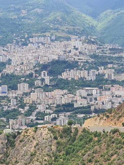 This is a wide aerial shot of the city of Artvin, Turkey, nestled in a mountainous, green landscape. The city is characterized by dense clusters of multi-story residential buildings with light-colored facades, many of which appear to be apartment blocks. The architecture is modern, with numerous balconies visible on the buildings. The city sprawls across the steep hillsides, with buildings built on terraced slopes.
In the foreground, a rugged, rocky terrain covered in sparse green vegetation slopes downwards. A road is visible winding up the hillside, with a few cars driving on it. Further up the hill, beyond the initial rocky slope, there are more clusters of buildings interspersed with trees. In the middle ground and background, the cityscape continues, with buildings appearing more densely packed as they recede into the distance. The entire scene is surrounded by lush green mountains and hills, indicating a high level of vegetation cover in the region. The sky is overcast, contributing to a muted lighting condition. Two yellow trucks are parked on a plateau on the hillside, near some buildings. No people are clearly discernible in the image.
