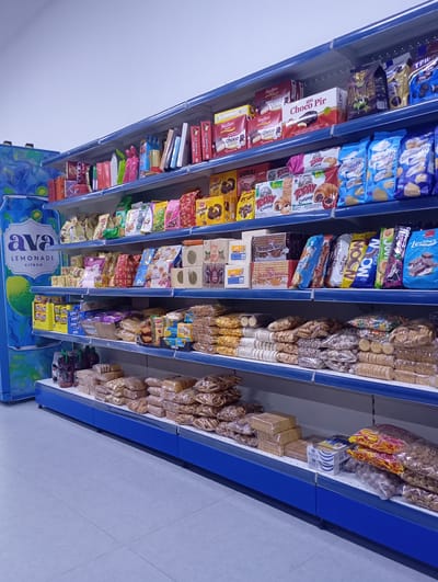 The image shows a well-stocked aisle in a supermarket or convenience store, specifically in the city of Arys, Kazakhstan. The shelves are filled with a variety of packaged food items, primarily sweets and snacks. Dominating the left side of the image is a large blue display panel featuring an advertisement for "Ava Lemonade Citron."
The shelves themselves are light blue and organized with numerous products. The top shelf displays boxes of cookies and chocolates, including brands like "Madlen Fabrica" and "Choco Pir." The second shelf down contains more packaged cookies and snacks, with colorful bags and boxes visible, such as "Dubai" and "Today Croissant" products, as well as several blue bags labeled with Russian text. The third shelf features more chocolate bars and cookies, including "Petit Beurre" and a product labeled "Gorilla." The bottom shelf displays an assortment of bagged snacks, cookies, and some bottled beverages on the far left. The floor is tiled with light gray squares. There are no people visible in the image. The overall impression is one of a typical retail environment.