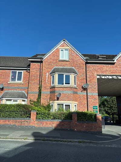 The image shows a row of red brick terraced houses with grey tiled roofs under a clear blue sky. The houses have white-framed windows and bay windows. There is a dark metal fence in front of a manicured hedge. A dark paved road runs in front of the fence, with a pavement to the left. A shadowed area from an unseen object falls across the road and pavement. To the right of the houses, an arched gateway leads to a driveway where a dark-colored car is parked. A green sign indicates "Gainsborough Court" with an arrow. The scene appears to be in a residential area in Crewe, United Kingdom.