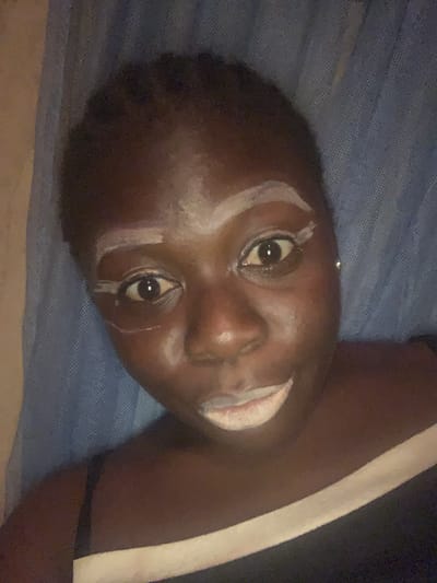 The image presents a close-up, rotated view of a dark-skinned individual, likely a child or young person, lying down and looking directly at the camera. The subject's face is adorned with prominent white markings: lines of a white substance are drawn above and below both eyes, extending onto the forehead in an arc, and also covering the area around the mouth. The eyes are wide open, engaging the viewer.
The person appears to be resting on a blue, textured fabric, which resembles a mosquito net or a patterned blanket, indicating an indoor setting. On the right side of the frame, a dark garment with a distinct white stripe is visible. The overall lighting is dim, suggesting that the photograph was taken in low light conditions, possibly during the evening or night. There is no visible text in the image.
Given the context of Kukum, Nigeria, the white facial markings are highly indicative of a traditional or cultural practice. Such body art is common in many Nigerian cultures for ceremonies, rituals, festivals, or as a form of traditional make-up, often using substances like chalk or clay. The image captures a moment of this cultural expression, with the individual presenting themselves directly to the camera.