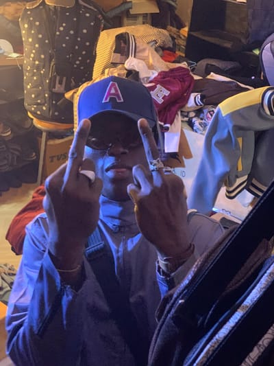 A young Black man is sitting in a cluttered room. He is wearing a blue jacket, a blue baseball cap with a white "A" on it, and a gold chain around his neck. He is making the middle finger gesture with both hands. The room is cluttered with clothes and other items. There is a wooden chair and a large backpack in the background. The location is in Ita Oniyan, Nigeria. The coordinates are 7.282 latitude and 5.161 longitude.