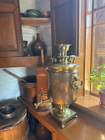 The image depicts an interior scene featuring a variety of antique household items, suggestive of a traditional Russian dwelling, possibly in Shushenskoye, Russia, given the context. In the foreground, a large, ornate brass samovar stands prominently on a wooden shelf. To the left of the samovar, a wooden bucket with a long handle is visible, and beside it, a tall wooden cylinder with a spigot. Further left, a wooden barrel with a lid sits on what appears to be a lower shelf or floor. Above these items, on a wooden shelf, are several ceramic pots, including a green one with painted designs and a reddish-brown jug. To the right of the samovar, a window with wooden panes is visible, and on the windowsill, a potted green plant. The overall impression is one of a preserved historical domestic setting. There are no people present in the image.