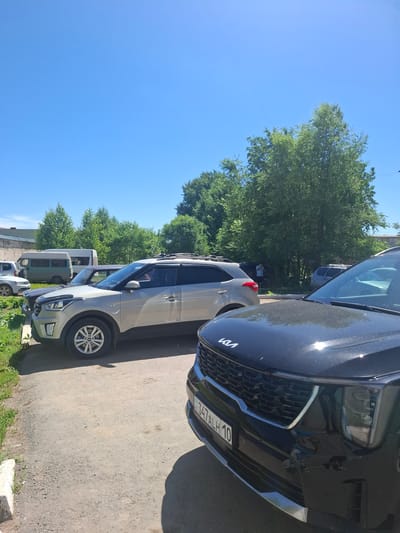 A black Kia SUV is parked in the foreground, facing right. The car's front grille, headlights, and license plate are visible. The license plate reads "G 347 ALH 10". Behind it, a silver SUV is parked, also facing right, with a roof rack. Further in the background, several trees and a light-colored van are visible. A person in a blue shirt is standing near the trees. The scene appears to be an outdoor parking area with a clear blue sky above and a paved surface. The location is identified as Jitiqara, Kazakhstan.