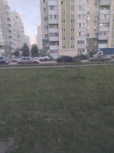 The image shows a street scene in a residential area. In the background are tall apartment buildings, characteristic of Soviet-era architecture, with visible balconies and windows. The buildings appear to be light-colored with green accents. A road runs in front of the buildings, and several cars are parked or driving on it. Among the cars, a light-colored sedan is prominent. To the left, another apartment building stretches into the distance. In the foreground, there is a grassy area, likely a lawn or park, with a small tree in the middle. The lighting suggests it is either early morning or late afternoon, with the sky appearing overcast. The overall atmosphere is quiet and typical of a suburban environment. There is no visible text in the image.
