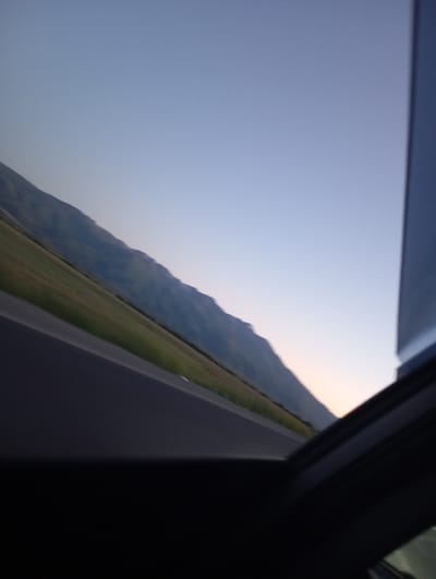 The image shows a view from inside a vehicle looking out at a landscape. The sky is a clear gradient of blue, transitioning to a lighter shade near the horizon. A mountain range runs diagonally across the frame, its slopes covered in green vegetation. In the foreground, a dark road runs parallel to the mountains. The image is taken from a slightly tilted perspective, giving a sense of motion. The scene appears to be set in the city of Kaysar, Kyrgyzstan, with the mountains suggesting a natural environment surrounding the urban area.