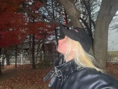 A woman with blonde hair and a black baseball cap is looking up. She is wearing a black leather jacket with silver buckles and zippers. The background includes a tree trunk, red-leaved trees, and a white fence surrounding a building. The ground is covered with fallen leaves. The lighting suggests it is dusk or a cloudy day.