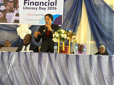 The image depicts an indoor event, likely a conference or seminar focused on financial literacy, set in Unguwan Dosa, Nigeria. In the foreground, a woman in a dark suit with a red top stands centrally, speaking into a microphone. She gestures with her left hand, wearing rings and a smartwatch.

Behind her, seated at a table draped in blue fabric, are several individuals. To the woman's left, a man in a dark pinstripe suit and tie is looking down, possibly at a laptop positioned on the table. Another man's face is partially visible to his left. To the speaker's right, a large floral arrangement with white, cream, and blue flowers sits on the table, flanked by two water bottles. Two women in hijabs, one purple and one black, are partially visible behind the floral arrangement. A cluster of white balloons is also visible on the table to the left of the speaker.

A large white banner dominates the background, announcing "Financial Literacy Day 2026" with the "Theme: Smart Mone" (likely "Smart Money"). The banner also includes contact information: "TOLL FREE HELP DESK: 080063424357," "HELP DESK E-MAIL," "WEBSITE," and social media handles for "@NDICNigeria" (Instagram, Facebook, and Twitter logos are implied). The word "Talks" is visible on the upper left of the banner. The backdrop features blue and white sheer curtains, behind which some text, including names like "YUSUN," "HALBU," "ARDA," and "SANDA," is faintly discernible on a board. The overall atmosphere suggests a formal educational or informational event.