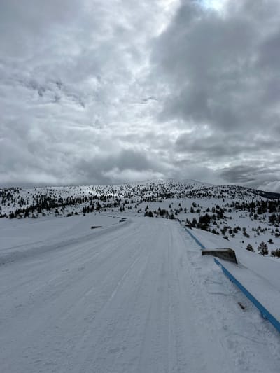 The image depicts a snow-covered landscape in Cherna Mesta, Bulgaria. A path cleared of snow runs diagonally across the frame. Trees line the left side of the path, extending up a mountain or hillside. A blue guardrail is visible along the right side of the path. Above the landscape, a cloudy, overcast sky fills the upper portion of the image. The scene is devoid of people or active subjects. The overall impression is that of a winter landscape with a maintained path.
