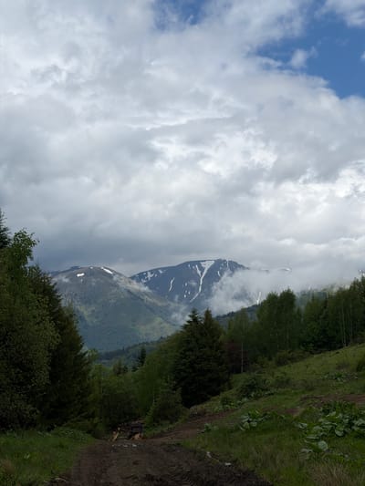 The image depicts a mountain range partially obscured by clouds, adjacent to a hillside path. Snow patches are visible on the mountain's slopes. The sky exhibits both blue patches and dense cloud cover. A dirt path with greenery borders the mountain view. The location is likely near Рилски манастир, Bulgaria, given the mountainous terrain and vegetation typical of the region.