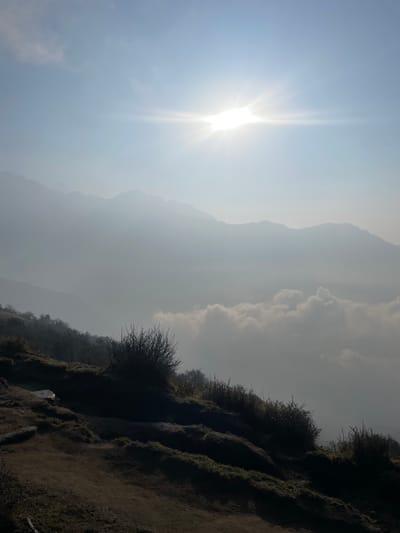 The image captures a mountain landscape under bright sunlight in Lwang Ghalel, Nepal. A prominent mountain range is visible in the background, obscured by haze. Clouds are dispersed among the mountains. In the foreground, the edge of a mountain or cliff is covered with sparse vegetation and soil. The bright sun dominates the left portion of the sky.