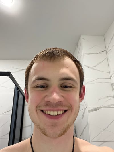 A smiling, bare-chested young adult male is depicted in a close-up, chest-up view. He has short, wet, light brown hair, a fair complexion with light stubble on his chin and jawline, and a minor reddish mark on his forehead. He wears a dark, possibly braided, necklace around his neck and smiles broadly, revealing his teeth. The background indicates an indoor, modern bathroom setting in the city of Tliustenkhabl', Russia, featuring rectangular wall tiles in white or light grey with subtle dark veining patterns. A plain white ceiling with a circular light fixture is visible above, and part of a dark-framed shower enclosure is present on the left side of the frame.