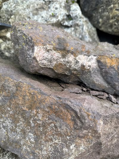 The scene presents a close-up, downward-angled view of a pile of large, irregularly shaped rocks. The dominant rocks in the foreground are dark grey to brown, exhibiting rough, weathered surfaces. Patches of white, yellow, and greenish lichen or mineral deposits are visible on these surfaces. A prominent upper rock partially overlaps a lower, flatter rock, creating a narrow, shadowed crevice. Within this crevice, several small, flat, dark-grey rock fragments or shale-like pieces are discernible. The texture of the rocks displays variations in color, including reddish-pink hues on some facets. The background consists of out-of-focus, similarly colored rocks. No individuals, animals, or man-made objects are present in the frame. The environment is consistent with an outdoor setting featuring natural rock formations. The depicted location is Krasnooktiabrskiy, Russia.