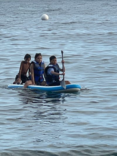 The image shows three young people on a paddleboard in the water, likely near Dauin, Philippines, as indicated by the presence of a spherical buoy floating in the distance, a common sight in coastal areas. The person in the front right is holding a paddle and appears to be in control of the board. They are wearing a blue life vest. To their left, another individual, also wearing a blue life vest, is seated. The third person, on the far left, is kneeling and appears to be shirtless. The water is a muted blue, with visible ripples indicating slight movement. The overall scene captures a moment of recreational activity on the water.