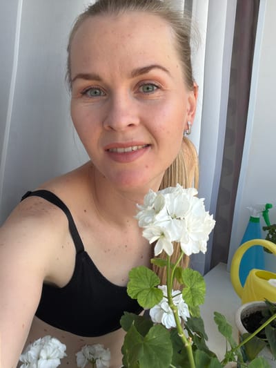 The image shows a close-up of a woman with blonde hair and blue-green eyes. She is wearing a black bra and smiling. She is holding a white flower in front of her face. In the background, there are white curtains, a yellow watering can, and a blue spray bottle. The scene appears to be indoors, possibly on a balcony, with natural light coming from the right. There is no information about the location being in the city of Кульдур, Russia.