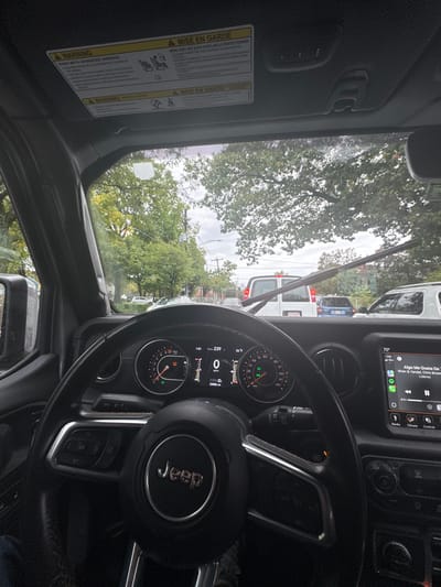 The image provides a driver's-eye view from within a black Jeep vehicle, stationary on a street in Newton, United States. The dashboard displays 0 MPH, an odometer reading of 23,900 miles, and an ambient temperature of 56°F. The steering wheel, featuring the "Jeep" logo, dominates the central foreground. To the right, an infotainment screen shows "Algo Me Gusta De Ti" by "Wisin & Yandel, Chris Brown." Overhead sun visor labels are visible in both English and French.
Through the windshield, a tree-lined street appears under an overcast sky, with the road surface appearing wet. A windshield wiper is positioned vertically, suggesting recent or active use. Traffic is visible ahead, including a white panel van immediately in front of the observer's vehicle, followed by a blue SUV/van. Green deciduous trees and utility poles line the street on both sides. A portion of a blue pant leg is visible in the lower-left corner of the driver's footwell area. The specific street or neighborhood within Newton is not discernible from the visual information.
