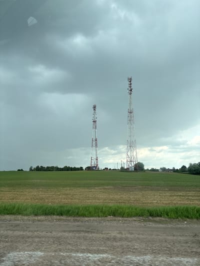 The image depicts a rural landscape in Голацкі сельскі Савет, Belarus. Two communication towers stand prominently against a cloudy, overcast sky. The towers are red and white lattice structures. A field of green and brown grass extends to the right of the towers. A dirt or gravel road runs along the right edge of the image. Trees and vegetation are visible in the distance. The light suggests it is daytime.