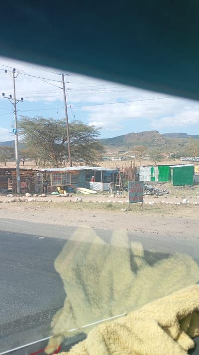 A roadside scene shows small businesses constructed from wood and corrugated metal. Power lines run along the road. A sign indicates car wash and parking services. A mountain rises in the distance under a partly cloudy sky. The view is partially obstructed by a dirty windshield with a visible reflection and a yellow blanket.