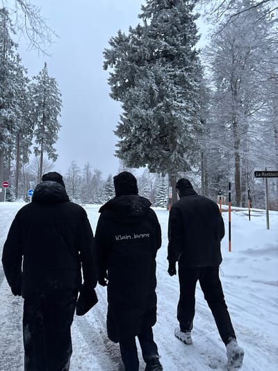 Three individuals walk away from the camera on a snow-covered path in Le Hohwald, France. The path is bordered by snow-laden evergreen and deciduous trees under an overcast sky. The person in the center wears a black winter coat with "Klein.jeans" visible in white text on the back. To their left, another individual wears a plain black winter coat, while the person on the right wears a black jacket and pants with white sneakers. On the left side of the path, a red circular 'no entry' sign and a blue circular 'mandatory right turn' sign are partially visible. On the right, a street sign reads "La Rothlach" and several orange and black striped poles stand upright in the snow. The ground is uniformly covered in snow, and tree branches are heavily frosted.