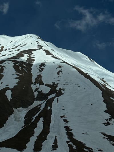 The image shows a steep mountain covered in snow with patches of dark rock or earth visible through the snow. The mountain is positioned against a clear blue sky with a few scattered clouds. The perspective is from below, looking upwards at the mountain's flank. There is no visible evidence of any people or structures. The location is assumed to be near Ganisi, Georgia, based on context, though this cannot be verified from the image alone.