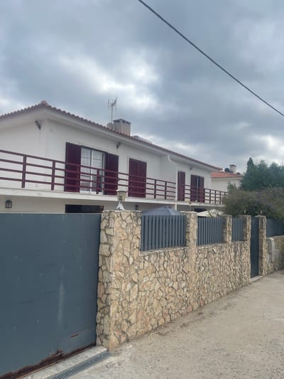 The image shows a residential property in São João das Lampas, Portugal, under an overcast sky. The property features a two-story white house with a red tile roof. The upper floor has a balcony with a maroon railing, and several windows with maroon shutters are visible. A chimney and an antenna are on the roof. In front of the house, a fence separates the property from the street. The fence is constructed of a stone wall base topped with dark gray vertical metal slats. A large, dark gray metal gate is on the left side of the image, partially obscuring the property entrance. The ground in front of the fence and gate is a dirt and gravel surface. Trees and bushes are visible behind the house on the right side.
