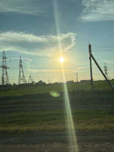 The image depicts a wide, rural landscape under a bright, hazy sky. The sun is a dominant element, positioned in the upper center of the frame and emitting a strong lens flare that creates a bright vertical beam across the scene. The sky is a pale blue, with wispy white clouds scattered towards the left. Power lines are a prominent feature, crisscrossing the frame horizontally and supported by tall, lattice-like towers.

In the midground, a vast, green field stretches across the landscape, suggesting agricultural use. Several large, dark green trucks or semi-trailer trucks are visible on a road or pathway running through the field. In the distance, a line of trees forms a dark, blurred horizon. The overall impression is one of a working landscape bathed in the warm glow of the setting or rising sun. No people are visible in the image. The location is identified as Vasilevka, Russia.