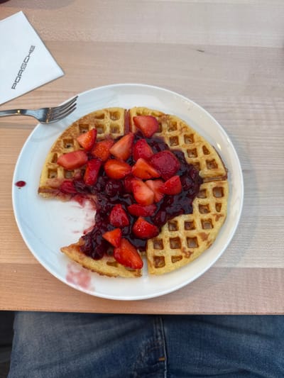 The image depicts a close-up, high-angle view of a partially consumed waffle presented on a white circular plate. The golden-brown waffle is topped with a generous portion of dark red berry compote and numerous sliced fresh strawberries. Approximately one-quarter of the waffle is missing from the left side, with residual red liquid stains visible on the white plate. A silver four-tine fork rests on the light-toned wooden table surface to the left of the plate. Above the fork, a folded white paper napkin displays the black "PORSCHE" logo printed vertically. The lower portion of the frame shows the upper legs of an individual wearing blue denim jeans, consistent with a first-person perspective from a seated position. The scene is located within the Porsche Centre Francorchamps in Stavelot, Belgium, indicated by the branded napkin.
