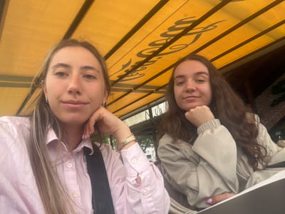 Two young women are taking a selfie outdoors, seated under a yellow awning. The woman on the left has long blonde hair and is wearing a pink striped button-down shirt. She is resting her chin on her hand. The woman on the right has long brown hair and is wearing a light-colored jacket. She is also resting her chin on her hand. The awning above them has a repeating logo or pattern in black. The background shows a building with brickwork and some greenery. The image appears to have been taken during the daytime, with natural light illuminating the scene.