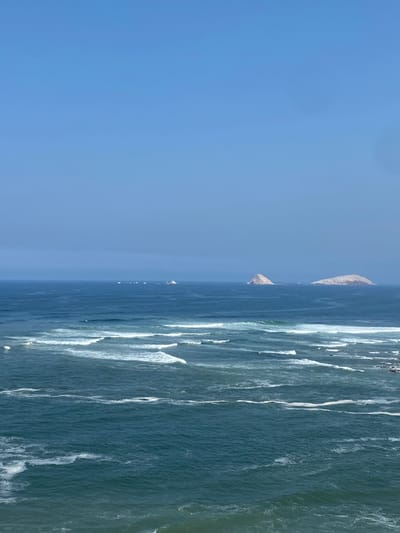 The image shows a view of the Pacific Ocean from the coast of Punta Hermosa, Peru, taken from coordinates -12.326 latitude and -76.834 longitude. The ocean is a deep blue with whitecaps from breaking waves in the distance. The horizon is a pale blue with no visible land masses except for three small islands in the distance. The sky is a clear, bright blue. There are no people or objects visible in the image.