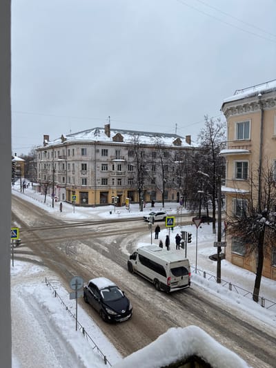 The scene depicts a snow-covered urban intersection in Velikiye Luki, Russia, specifically at the corner of Komsomolskaya Ulitsa and Ulitsa Karla Libknekhta, viewed from an elevated position, likely a building balcony. The sky is overcast and grey.
The ground is blanketed in snow. Roadways are partially cleared, revealing wet, dark asphalt and slush, with visible tire tracks. Sidewalks and unplowed areas are covered in a deeper layer of white snow. Bare deciduous trees with snow clinging to their branches are interspersed throughout the environment.
In the midground-left, a multi-story (at least four floors) light-colored building, primarily off-white or pale beige, with yellow-painted commercial facades on its ground floor, forms the prominent corner of the intersection, identified as Komsomolskaya Ulitsa 29 / Ulitsa Karla Libknekhta 12. Its roof and ledges are covered in snow, and it features classical architectural elements including cornices and balconies. On the right, a section of a similar light-yellow building, likely Komsomolskaya Ulitsa 31, is visible with snow and icicles on its roof and decorative moldings.
Vehicle activity includes a black Peugeot 308 sedan, with snow accumulated on its roof and hood, traversing the foreground from left to right. Its Russian license plate is partially visible. A white commercial van or minibus is positioned in the midground, also moving left to right, near a group of pedestrians. A white SUV is stationary at a crosswalk further up the street on the left.
Four pedestrians, dressed in winter clothing, are standing on the snow-covered sidewalk in the midground-right, adjacent to a traffic light and a pedestrian crossing, appearing to wait to cross the street. Additional individuals are visible walking on sidewalks in the background.
Visible road signs include multiple blue-and-white pedestrian crossing signs, a red-and-white "СТОП" (STOP) sign mounted on a pole, and a "Give Way" sign. Operational traffic lights are also present. The immediate foreground shows a snow-covered balcony railing, indicating the elevated viewpoint. Overhead power lines span the sky.