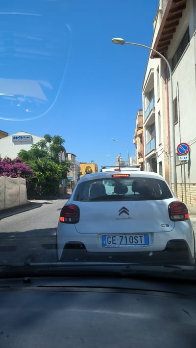 The image shows a rear view of a white Citroen C3 car with the license plate "GE 710ST" in Cinisi, Italy. The car is on a street with buildings on the right and trees and a building with a "WARM IMPIANTI" sign on the left. The sky is clear blue. There is a street light visible in the sky and a "No Parking" sign on the right side of the street. The scene appears to be a sunny day.
