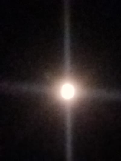 The image shows a bright, luminous circle in the center of a dark sky, resembling the moon or a distant light source. A distinct cross-shaped illumination emanates from this central point, extending outwards into the darkness. The light is diffused, suggesting it might be passing through atmospheric conditions like fog or dust. The overall impression is of a celestial event or a powerful light piercing the night. There are no discernible people, objects, or specific location cues within the frame that would indicate a particular setting beyond the dark sky. The time of day is clearly night due to the darkness. The weather is not definitively discernible, though the diffusion of light could suggest overcast conditions or the presence of moisture. No text is visible in the image.