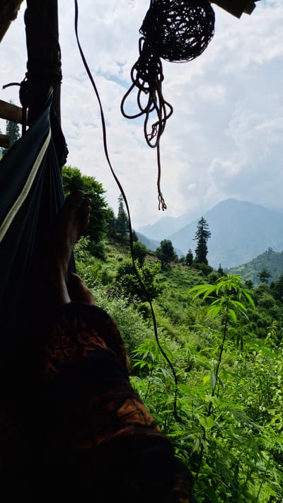 The image shows a person lying in a hammock, viewed from a low angle. The person's legs and feet are visible, partially obscured by dark fabric with a patterned trim. The hammock itself is dark blue with a lighter trim. The scene is outdoors, with lush green vegetation and rolling hills or mountains in the background. The sky is visible above, with some clouds. There are also ropes and a dark, spherical object hanging near the hammock, which appear to be part of the hammock's suspension or an adjacent structure. The overall impression is one of relaxation in a natural, possibly mountainous, setting. The lighting suggests it is daytime, possibly late afternoon given the shadows. The location cues, such as the dense greenery and mountainous terrain, are consistent with a place like Shila, India, known for its natural beauty and trekking opportunities. No visible text is present in the image.