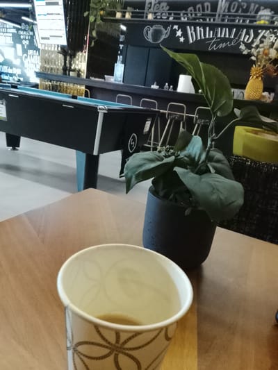The image depicts an indoor scene, likely a cafe, lounge, or entertainment area, possibly in Birkirkara, Malta. In the immediate foreground, a used paper coffee cup with a geometric pattern sits on a light wooden table, containing a small amount of leftover beverage. To the right of the cup, on the same table, is a potted plant with broad green leaves in a dark grey pot.
In the midground, a black pool table with a light blue playing surface is prominent, suggesting recreational activities are available. Behind the pool table is a bar or counter area with several dark-colored stools tucked underneath. The wall behind the counter is dark and features white chalk-style drawings and text.
Visible text on the wall includes "Tea," "Good Morn" (likely "Good Morning"), and "BREAKFAST time," accompanied by a drawing of a teapot, indicating the establishment serves refreshments and meals. Further to the left, more text reads "CARDS, IM ALL IN PLAYING" and "LIFE IS A GAMBL" (presumably "LIFE IS A GAMBLE"), alongside what appears to be a list or menu. Decorative elements like a golden pineapple are also visible on the counter. The overall setting suggests a casual and social environment, possibly frequented during the daytime given the "Breakfast time" cue. No people are present in the frame.