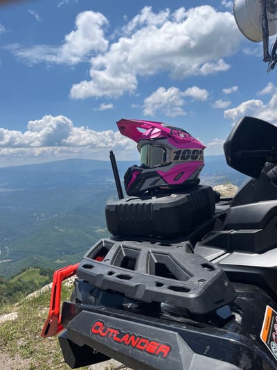In Simeonovets, Bulgaria, a pink and gray motocross helmet with goggles rests on the rear rack of a black Can-Am Outlander ATV. The ATV is parked on a rocky, grassy mountain overlook, with a vast, green, mountainous landscape stretching into the distance under a bright blue sky filled with white, fluffy clouds. A large white satellite dish is visible on a pole in the upper right corner of the frame.